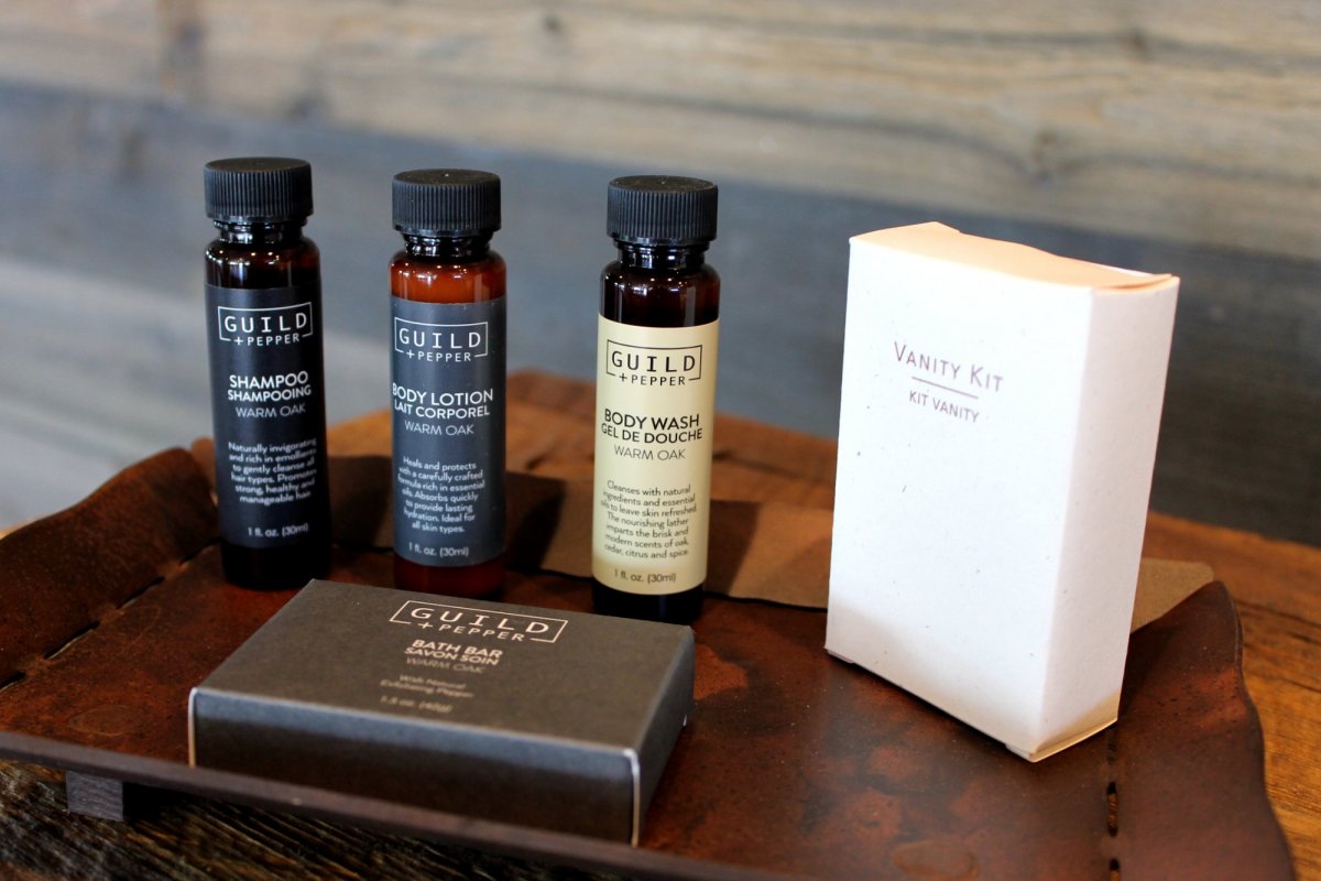 A brown tray holds 1872 Inn West Yellowstone amenities like Guild+Pepper shampoo, body lotion, body wash, a vanity kit box, and a bath bar, all arranged neatly on a wooden surface.